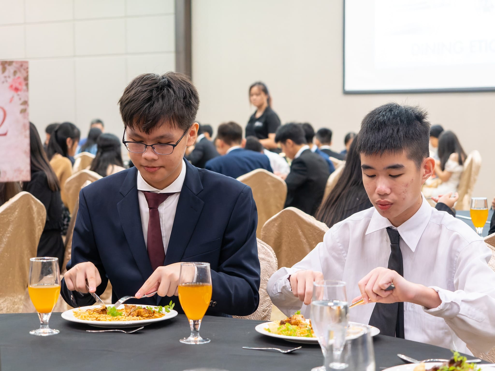 Two young men in formal attire eat rice dishes with orange juice at a banquet hall filled with seated guests.